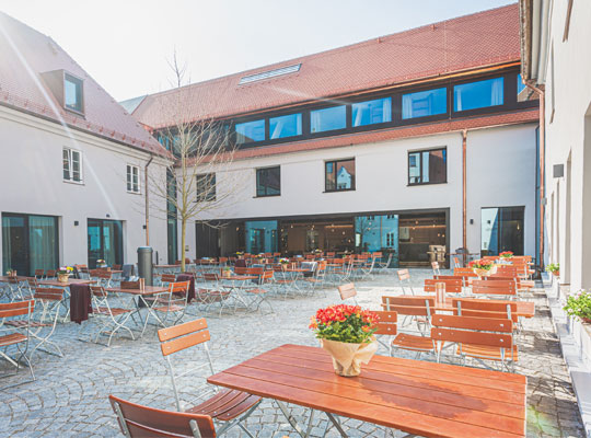 Auf der Terrasse stehen braune Holzstühle und Holztische und die Terrassentür vom Restaurant steht offen.